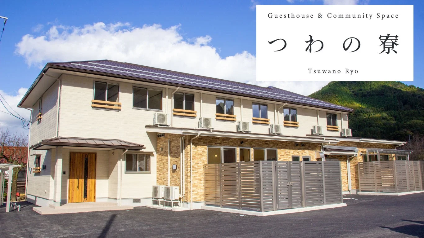 青空の下、モダンな2階建てのゲストハウス「つわの寮」の外観が写っています。奥には緑豊かな山が見えます。