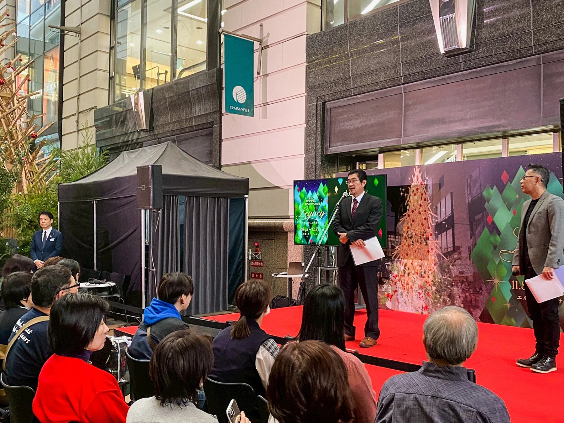 大丸百貨店で開催されたクリスマスイベントの様子です。ステージでは二人の男性が話し、多くの観客が熱心に聞き入っています。