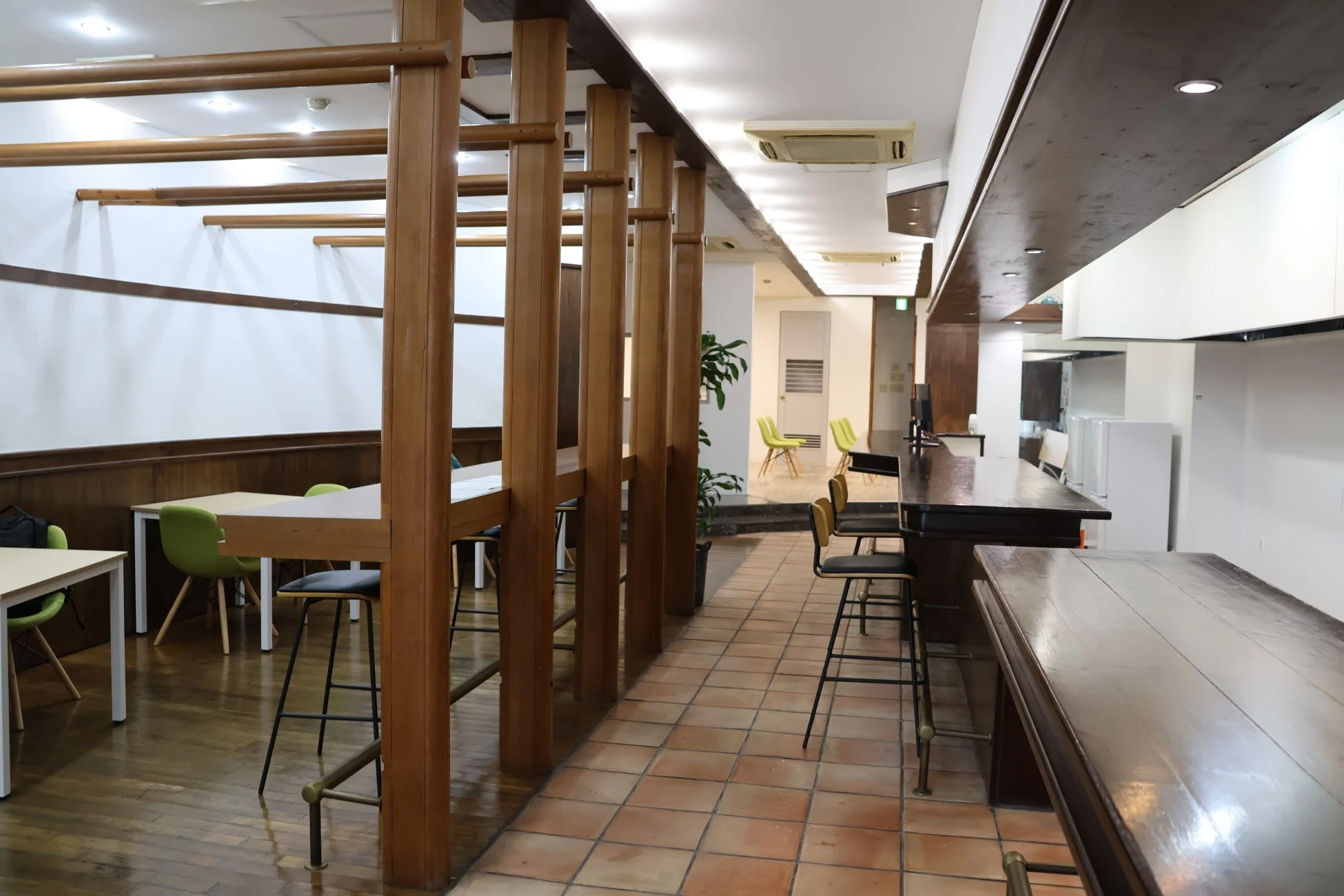 木の温もりを感じるモダンな室内空間。左は作業スペースで明るいテーブルと椅子、右はカウンター席とキッチンエリアがあり、多様な利用が想定される。