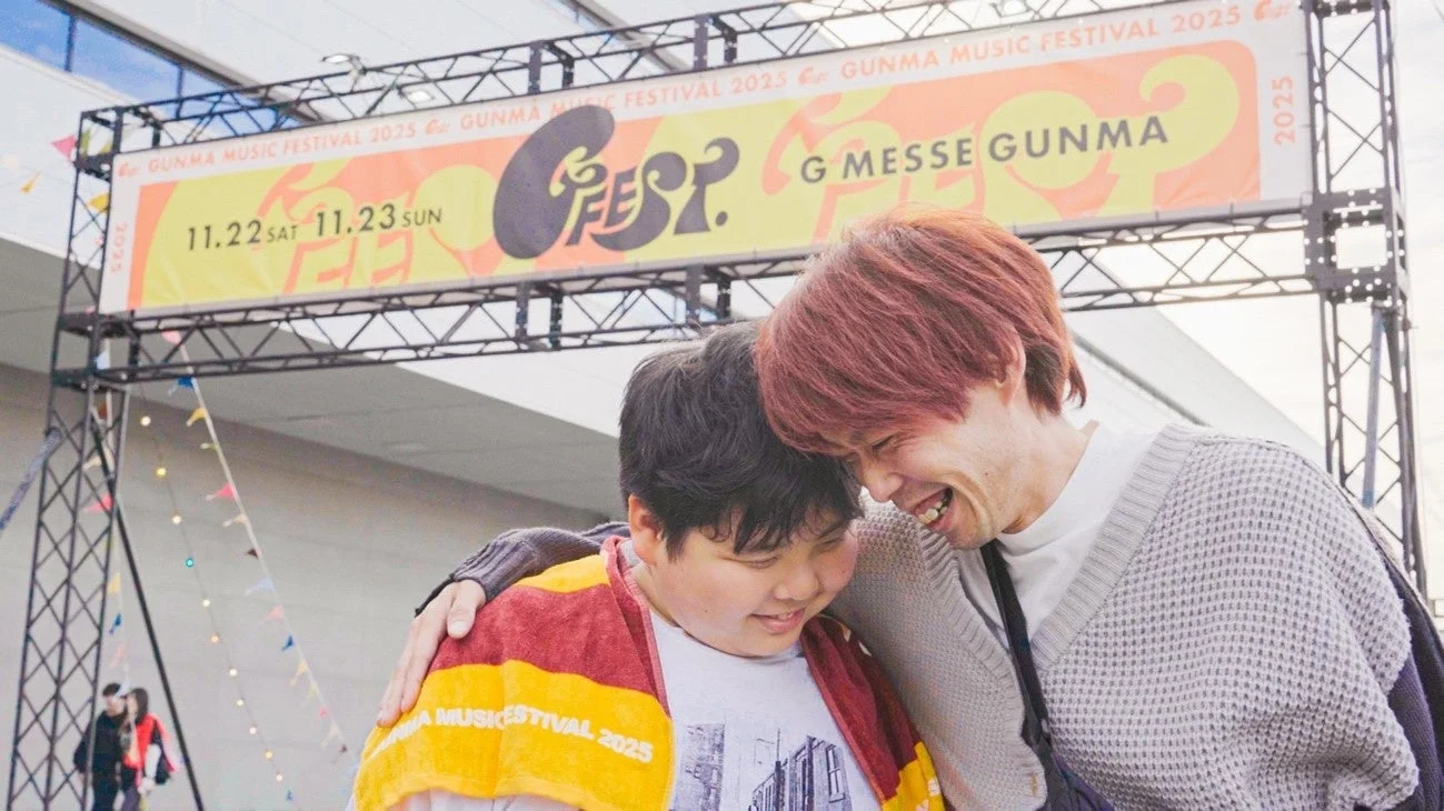 Gメッセ群馬で開催される「GUNMA MUSIC FESTIVAL 2025」のバナーを背景に、楽しそうに笑い合う2人の男性が抱き合っている瞬間を捉えた画像です。一人はフェスタオルを肩にかけています。和やかな雰囲気が伝わってきます。