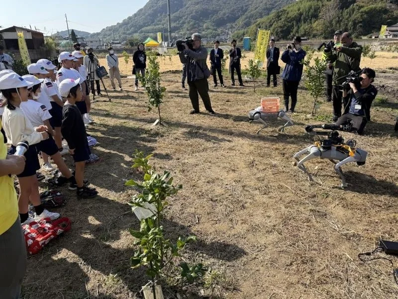 畑で、子供たちとカメラマンが見守る中、2台の四足歩行ロボットがデモンストレーションを行っている様子。農業分野でのロボット活用イベントと思われる。