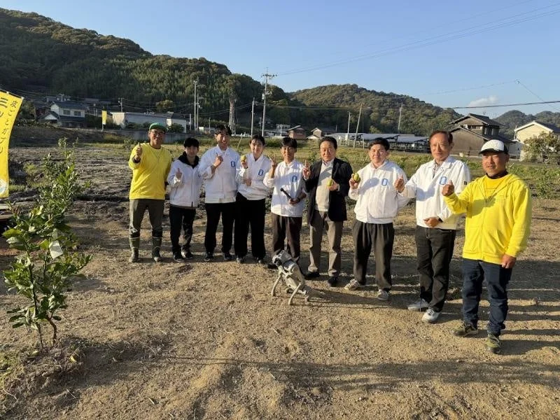 畑で10人ほどのグループと1匹の犬がポーズをとっている。参加者の多くは白いジャケットを着ており、数人が緑色の果物を持っている。植樹や収穫など、農業に関連するイベントの様子を捉えた写真である。