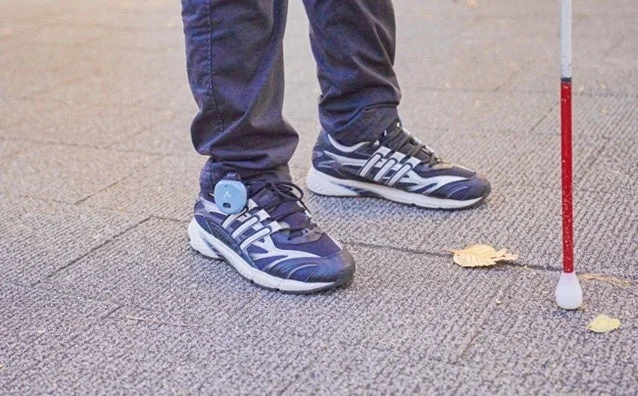 視覚障害を持つ人が白杖と、靴に装着された青い補助デバイスを使って歩いている足元のクローズアップ。屋外の地面に落ち葉が散っている様子が写されている。