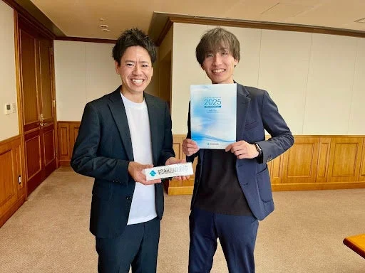 ビジネススーツ姿の男性二人が笑顔で立っており、一人は「特別優待」と書かれた箱を、もう一人は「2025」と書かれた書類を持っています。