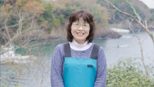 青いエプロンを着て笑顔で立つ中年の女性