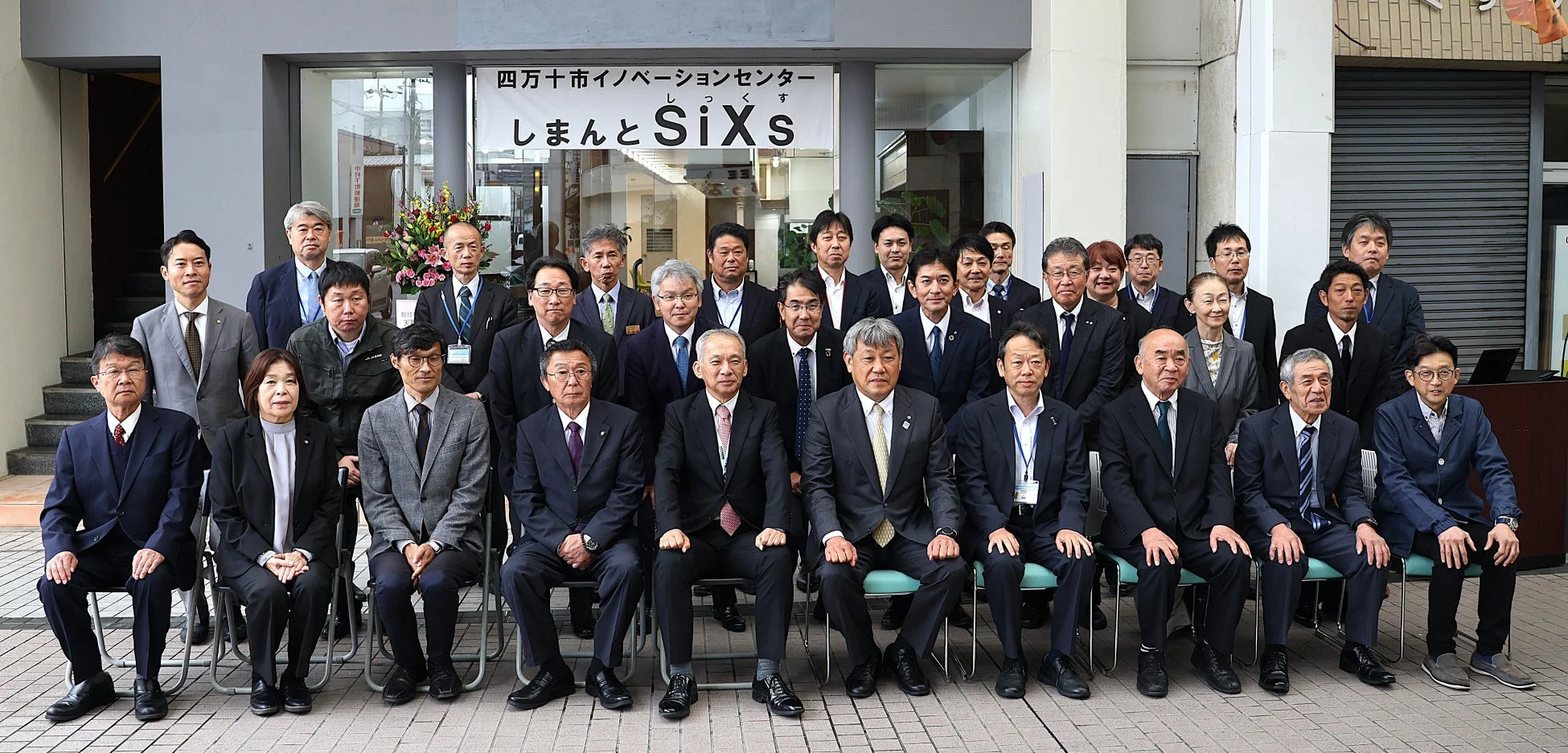 四万十市イノベーションセンター「しまんとSiXs」の前で撮影された、多数のビジネスパーソンによる集合写真です。スーツ姿の男女が整列し、カメラに向かってポーズをとっています。