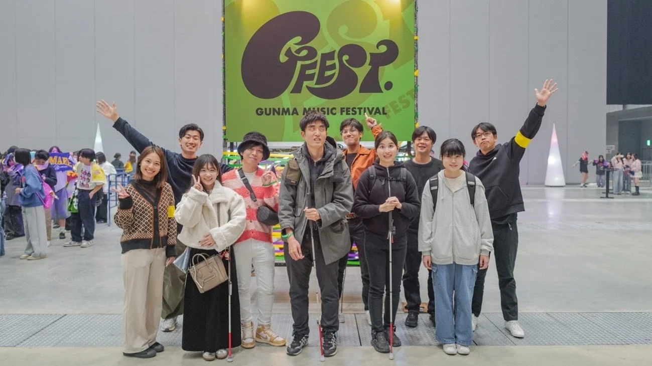 群馬音楽フェスティバル「GFEST.」のバナー前で、笑顔でポーズをとるグループの人々。白杖を持つ参加者もおり、イベントの多様性と楽しげな雰囲気が伝わります。