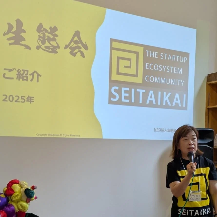 「生態会（SEITAIKAI）」のイベントで、一人の女性がマイクを持ってプレゼンテーションを行っている