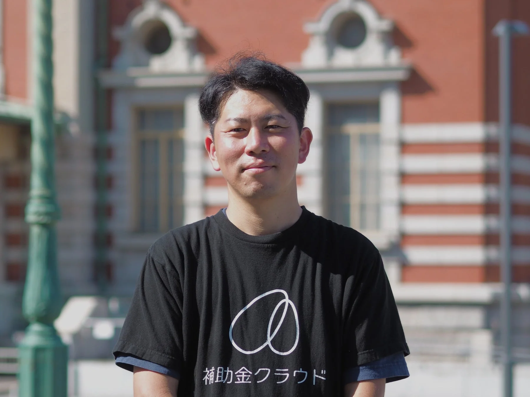 黒いTシャツを着た男性が屋外でカメラに向かって微笑んでいる。背景には赤レンガ調の建物が見える。