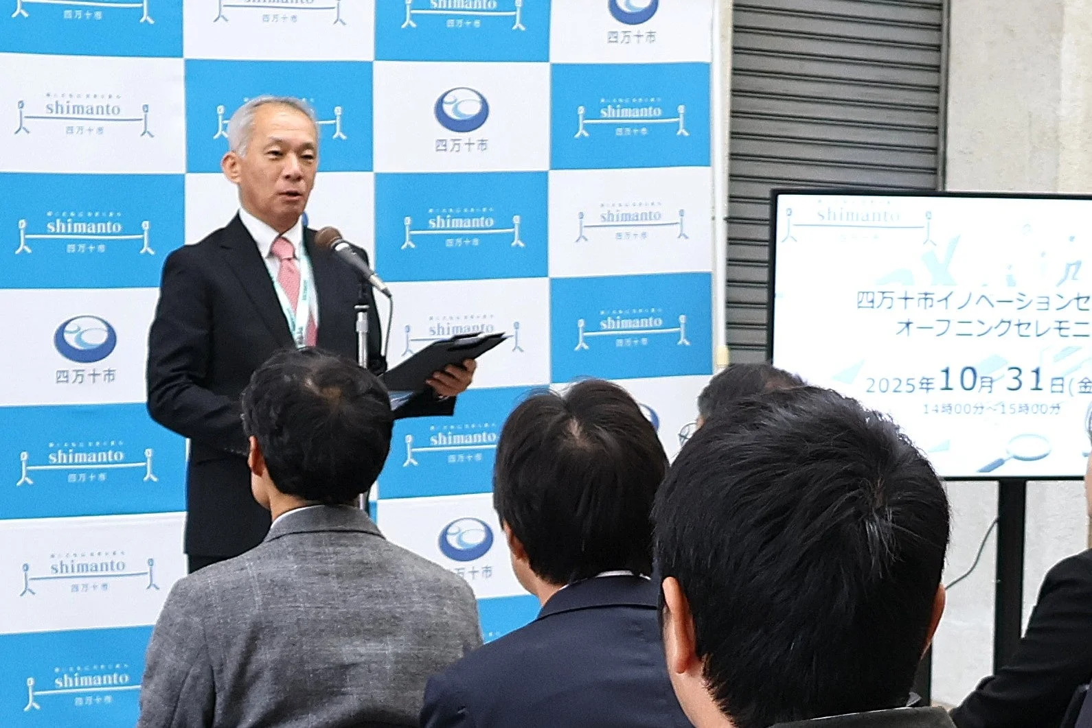 四万十市イノベーションセンターのオープニングセレモニーで、スーツを着た男性が講演しています。聴衆が耳を傾け、スクリーンには開催日時が表示されています。