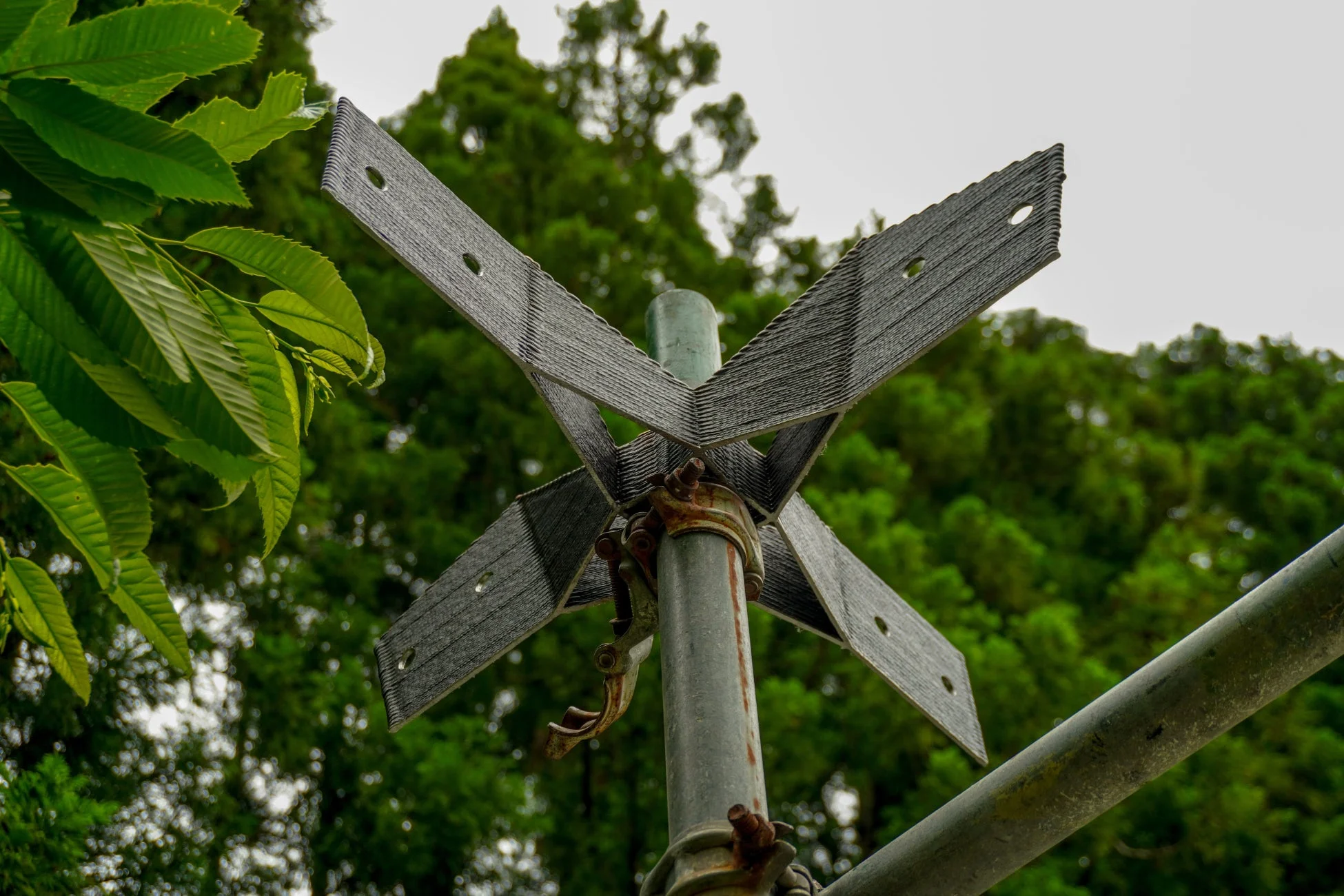 金属製のクロス状の構造物が支柱に取り付けられており、手前には緑の葉、背景には多くの木々が見える屋外の風景