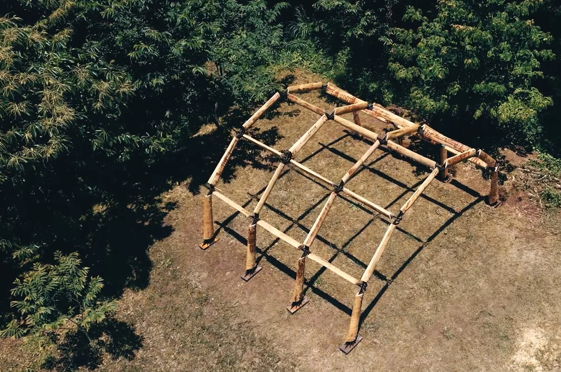 緑豊かな自然の中に設置された、シンプルな木造の建築フレームを上空から捉えた写真
