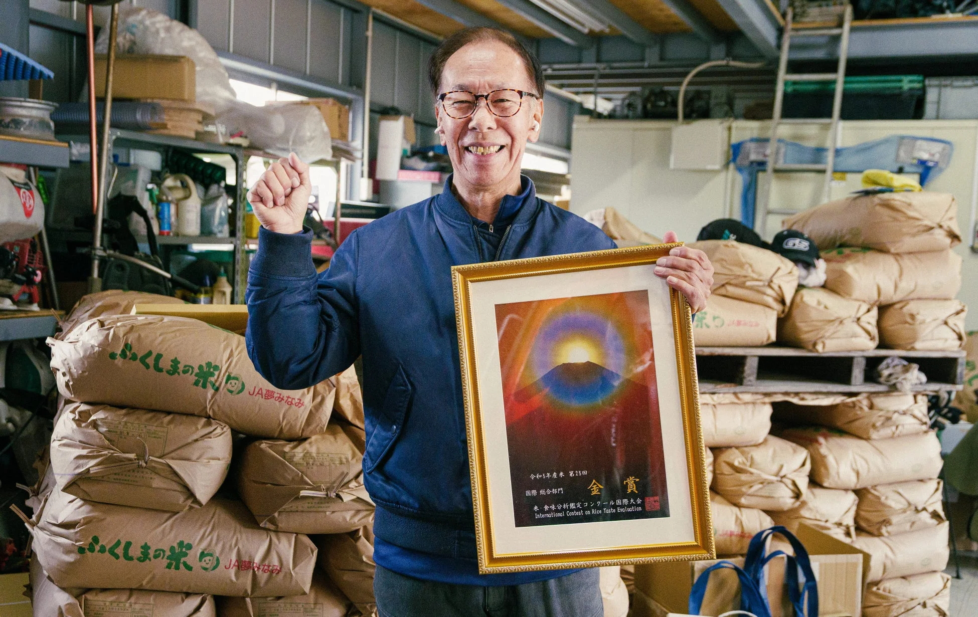 米袋が積まれた倉庫で、男性が「米・食味分析鑑定コンクール国際大会」の金賞状を掲げ、誇らしげに笑顔を見せています