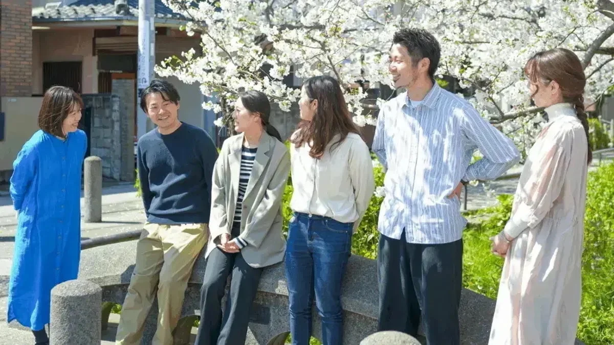 満開の桜の下、6人の男女が和やかに会話を楽しんでいます。