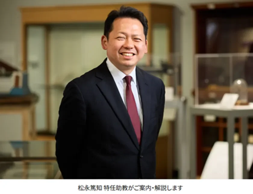 スーツとネクタイを着用した笑顔の男性がカメラに向かって立っています。背景には展示ケースのようなものが写っており、屋内で撮影された松永篤知特任助教のポートレートです。