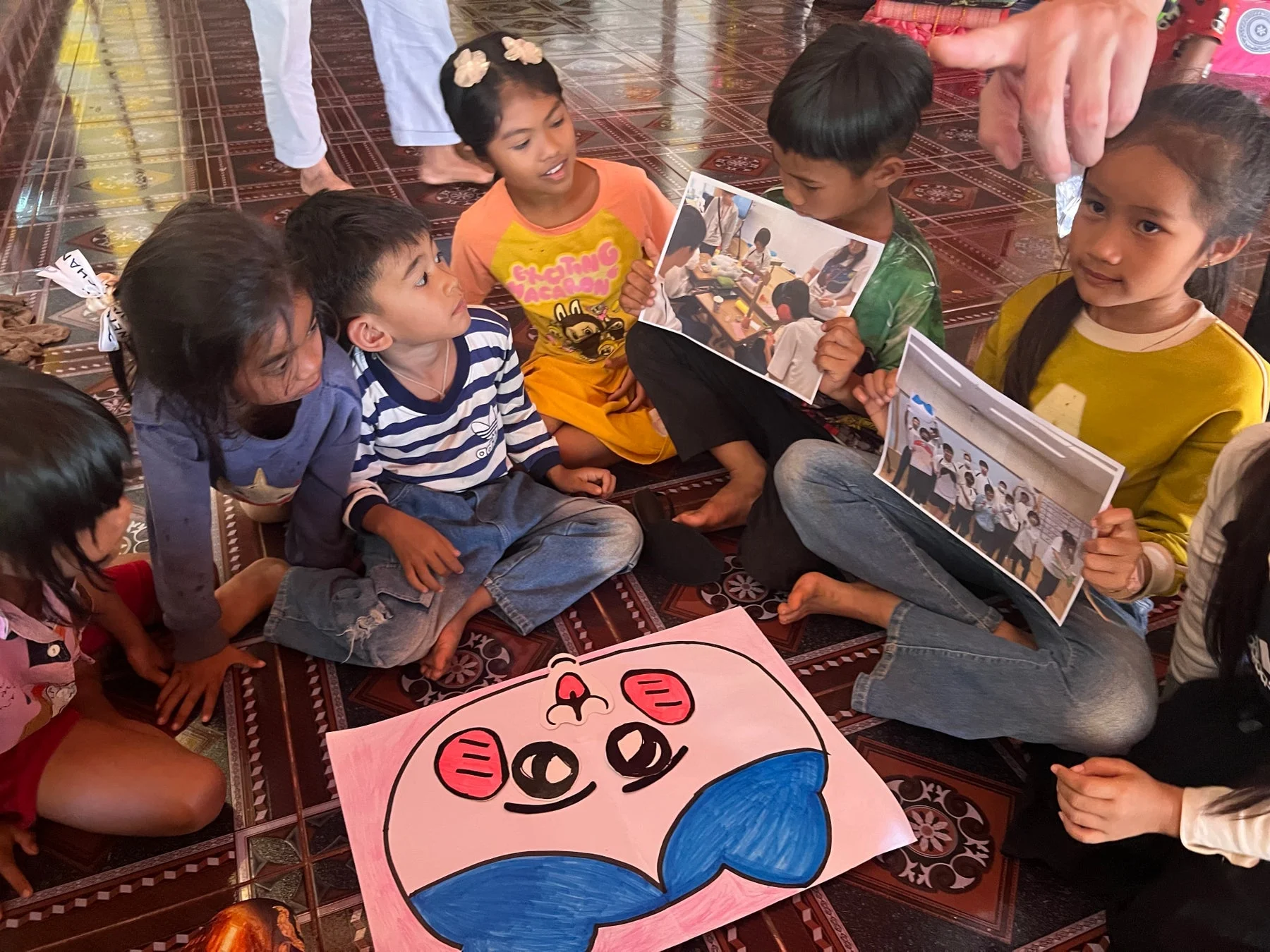 複数の子供たちが床に座り、写真を見せ合ったり、大きなイラストを囲んだりして交流している様子です。