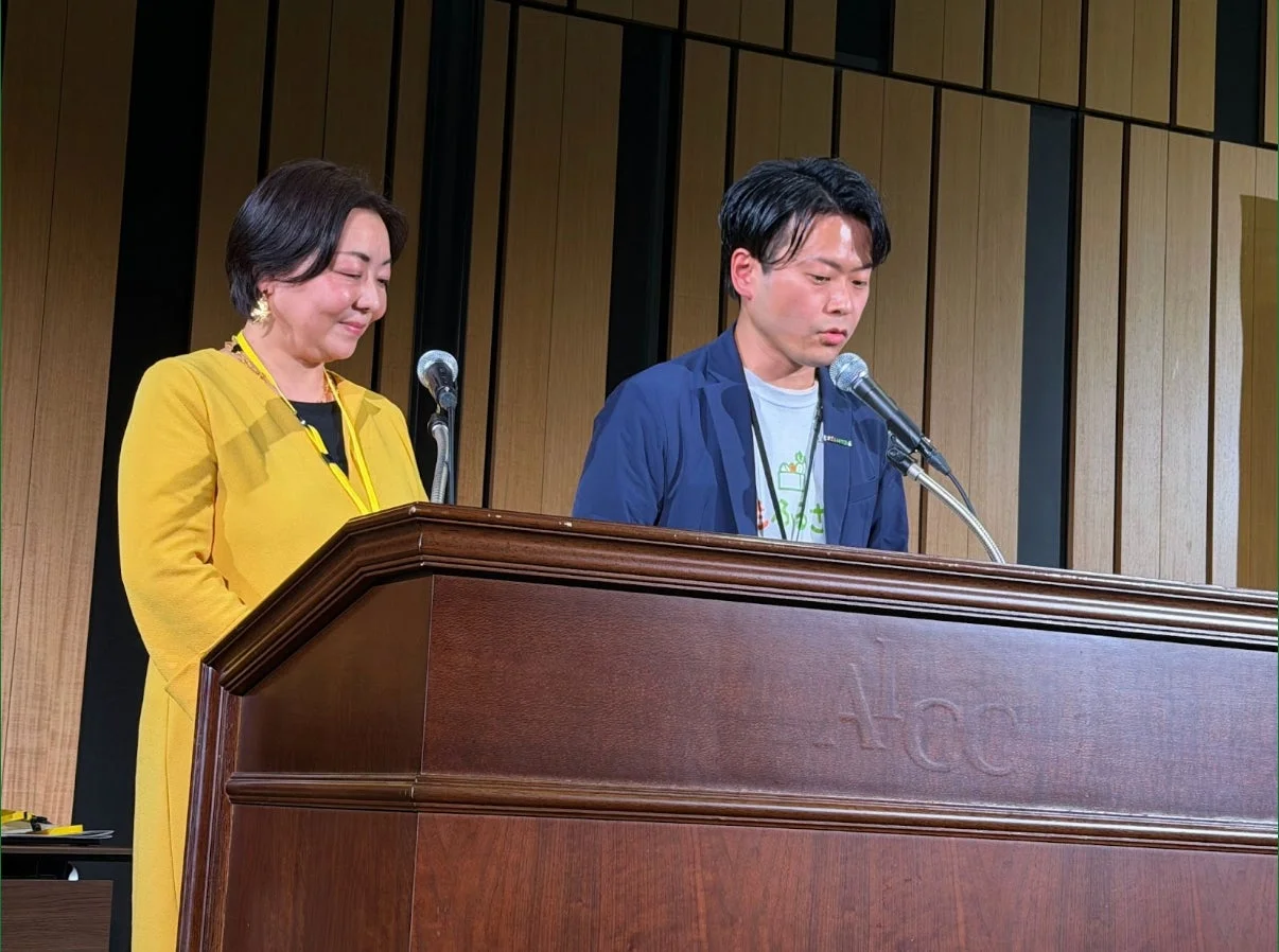 講演会らしき場所で、女性と男性の二人が木製の演台の前に立っています。男性はマイクを使って話しており、女性は隣で静かに見守っています。