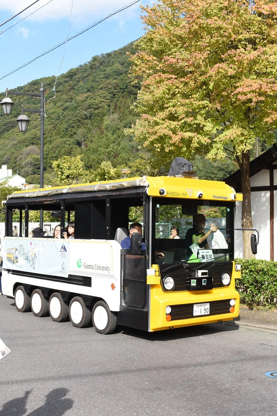 群馬大学のロゴが入った電気バスが紅葉の道を走行