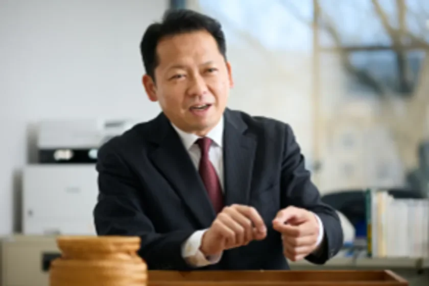 スーツ姿のアジア人男性がオフィスらしき場所で、ジェスチャーを交えながら話しているポートレート。真剣な表情で何かを説明している様子が伺えます。