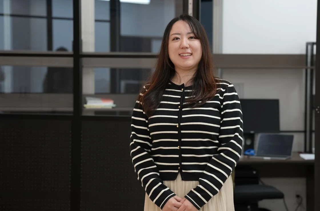 オフィスのような室内で、黒と白のストライプのカーディガンを着た笑顔の若い女性