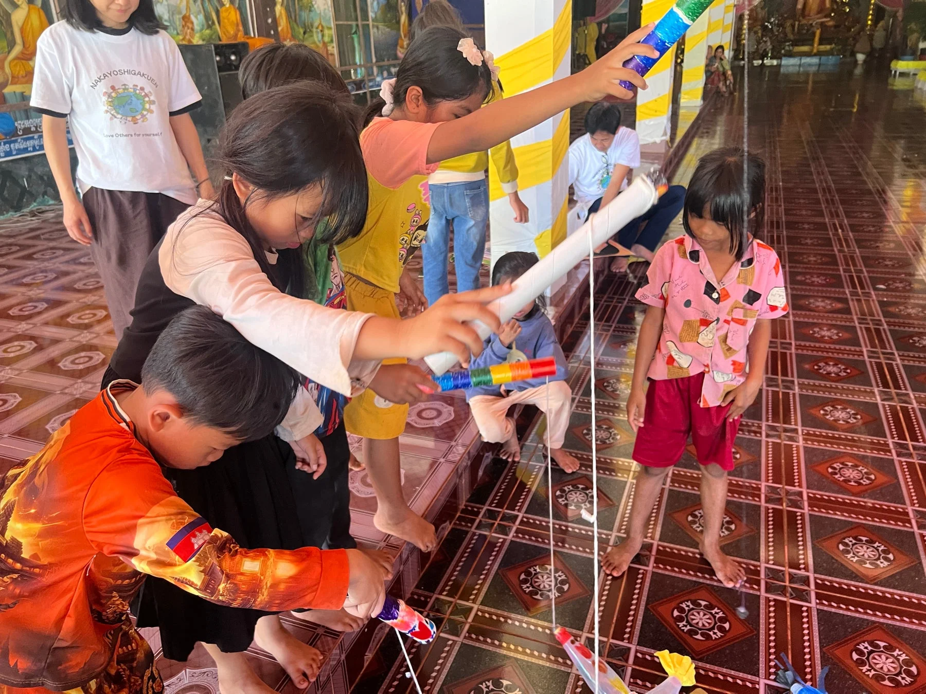 寺院のような場所で、複数の子供たちが集まり、ひもを使った手作りのおもちゃや道具で遊んでいる様子です。