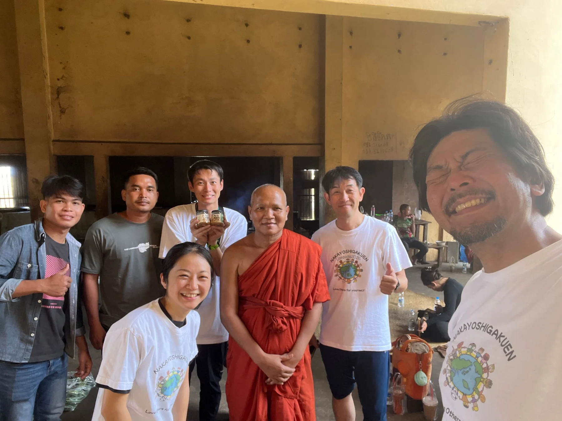 カンボジアの屋内と思われる場所で、僧侶と数名の男女が笑顔で写るグループ写真。参加者は「NAKAYOSHIGAKUEN Cambodia 2026」のTシャツを着用しています。