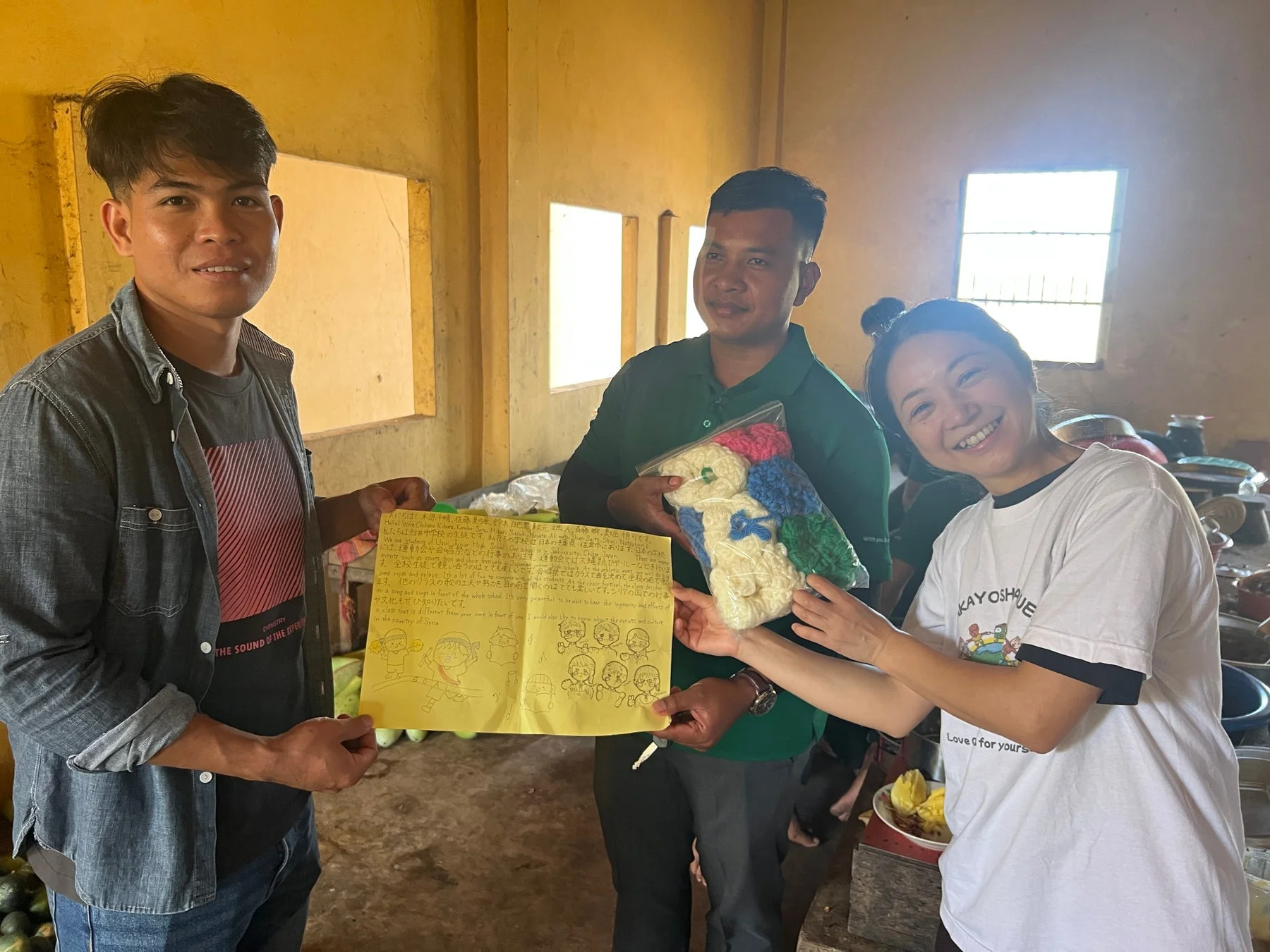 室内で3人の人物が交流している様子。一人が日本語と英語で書かれた手紙を持ち、もう二人がカラフルな編み物を交換しています。