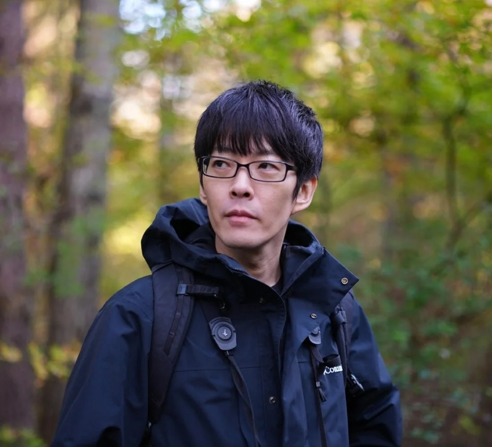 森の中で紺色のジャケットを着て眼鏡をかけた男性のポートレート。男性は少し上を見上げており、背景にはぼやけた緑と黄色の木々が広がる。自然の中での思索的な雰囲気が漂う一枚。