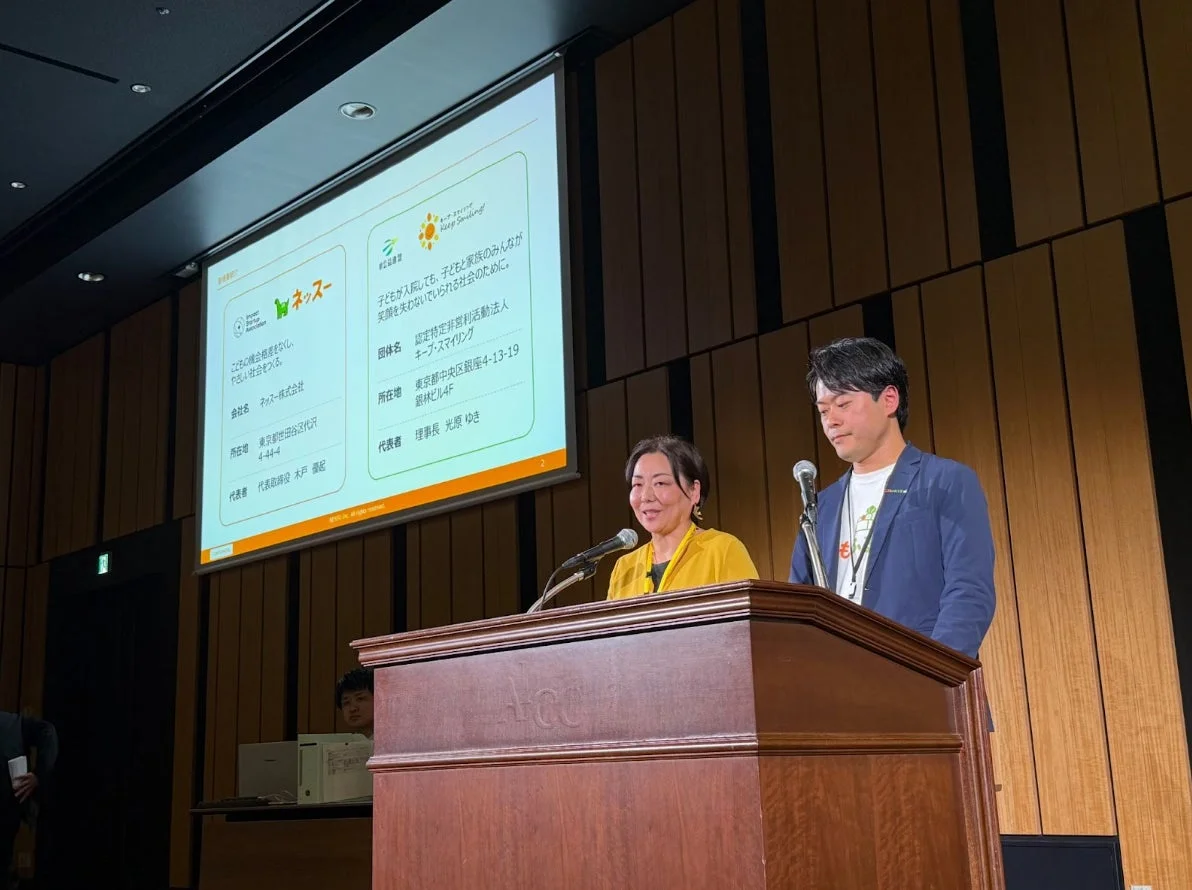 イベントで講演する男女2名の登壇者が写っています。背後のスクリーンには、こども縁組を促す「ネッズー」と、子どもと家族の笑顔を支援する「認定特定非営利活動法人 キープ・スマイリング」についての詳細な情報が映し出されています。