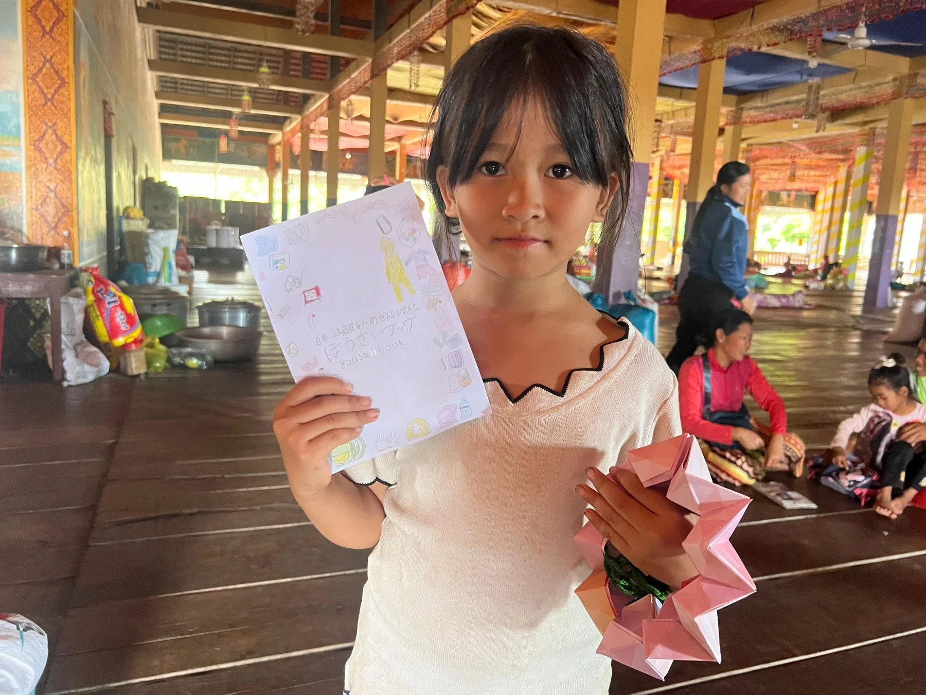 一人の少女が「ぼうさいブック」とピンク色の折り紙を持ってカメラを見ています。