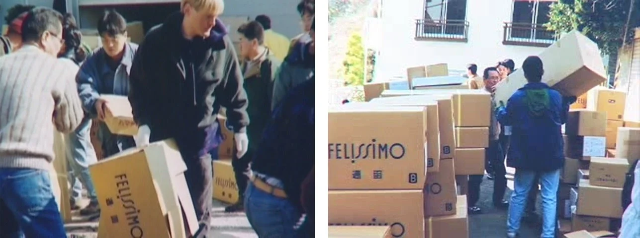 多くの人々が屋外で段ボール箱を運んだり積み重ねたりしている様子。箱には「FELISSIMO」のロゴが見られ、大量の荷物の配送や仕分け作業を行っていると思われる。