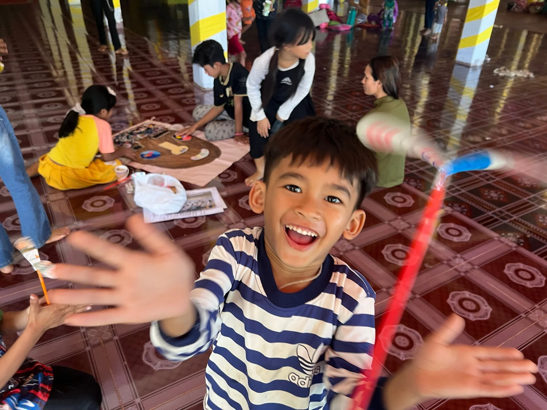 縞模様のシャツを着た少年が満面の笑みでカメラを見ており、他の子供たちは床に座って絵を描く活動に没頭しています。