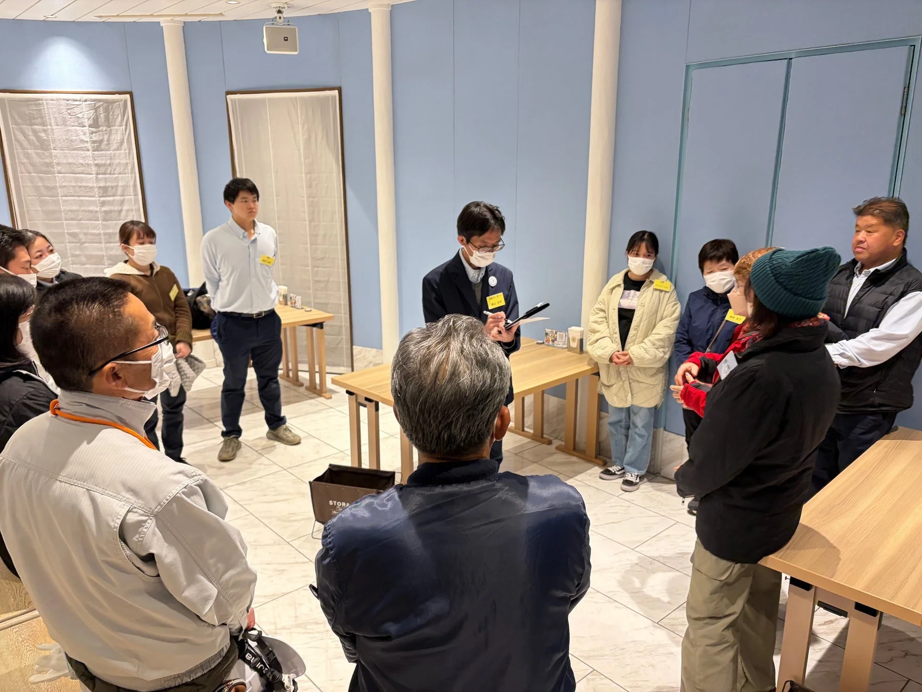 水色の壁の部屋に多くの人々が集まり、マスクを着用している様子