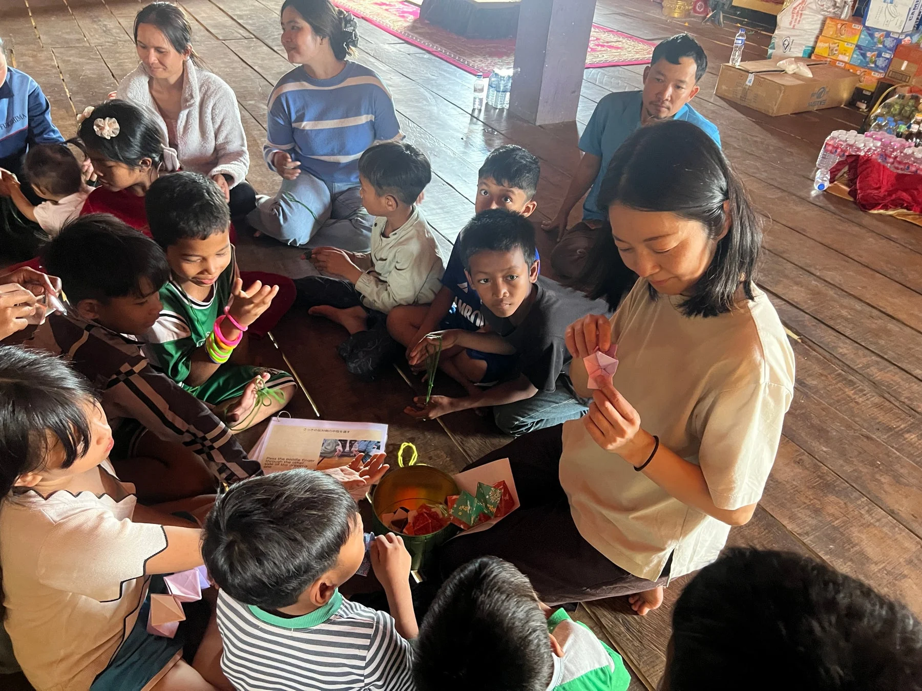 木の床に座り、折り紙のようなクラフト活動に熱中する子供たちと大人たちの様子です。