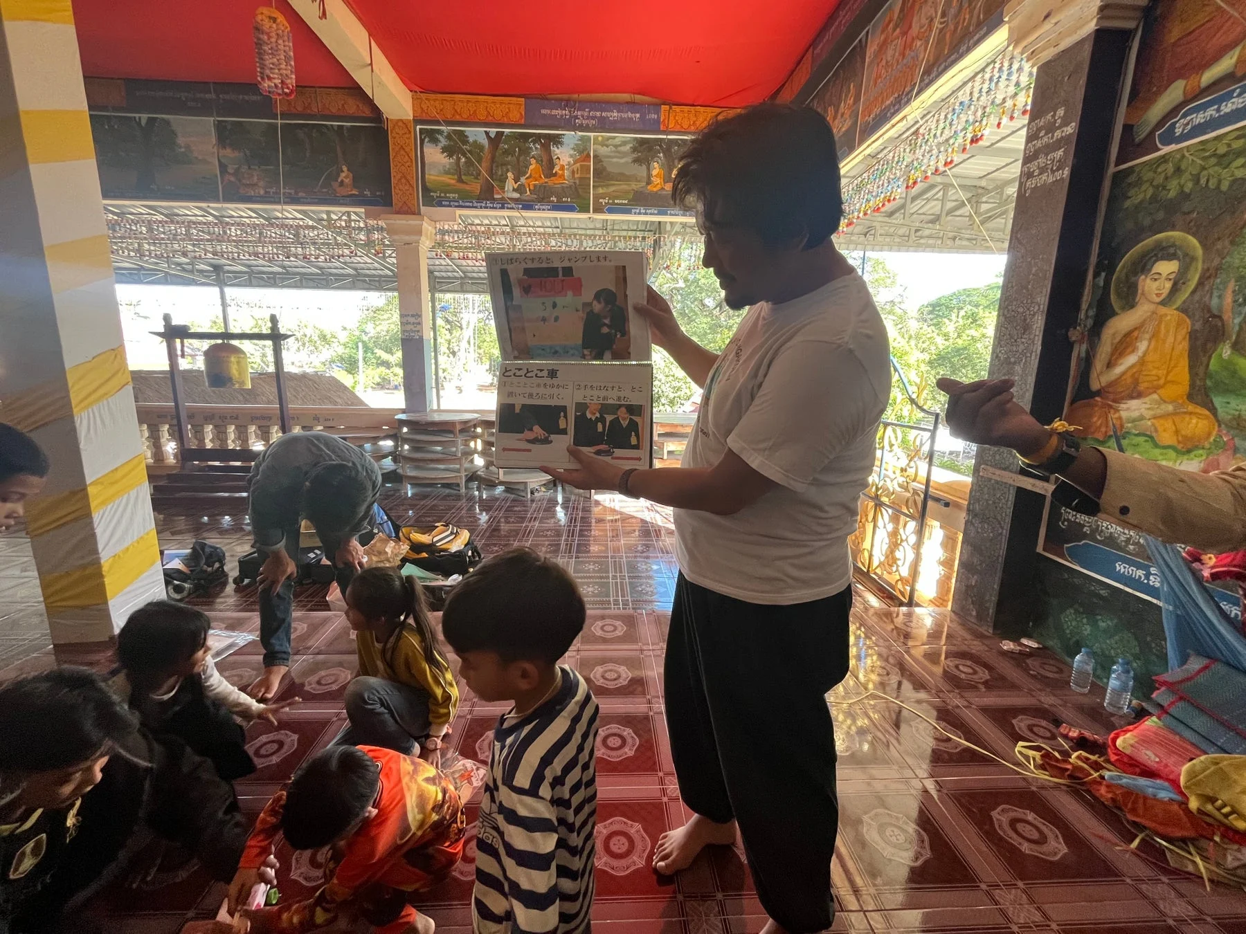 寺院のような場所で、男性が子供たちに日本語の教材を見せて説明しています。
