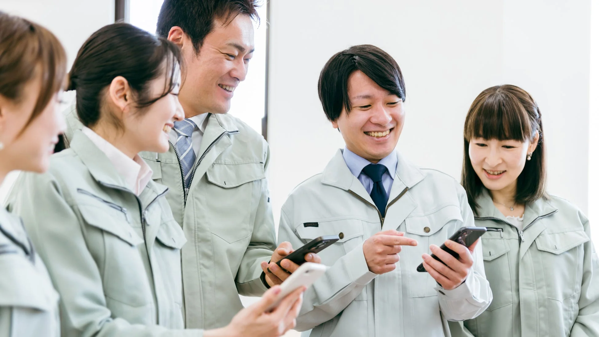作業着を着た男女5人がスマートフォンを見ながら笑顔で会話
