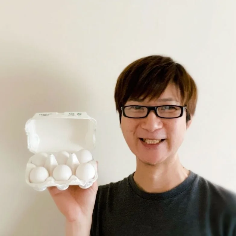 眼鏡をかけた男性が笑顔でカメラを見ており、片手に卵が6個入った白い卵パックを持っています