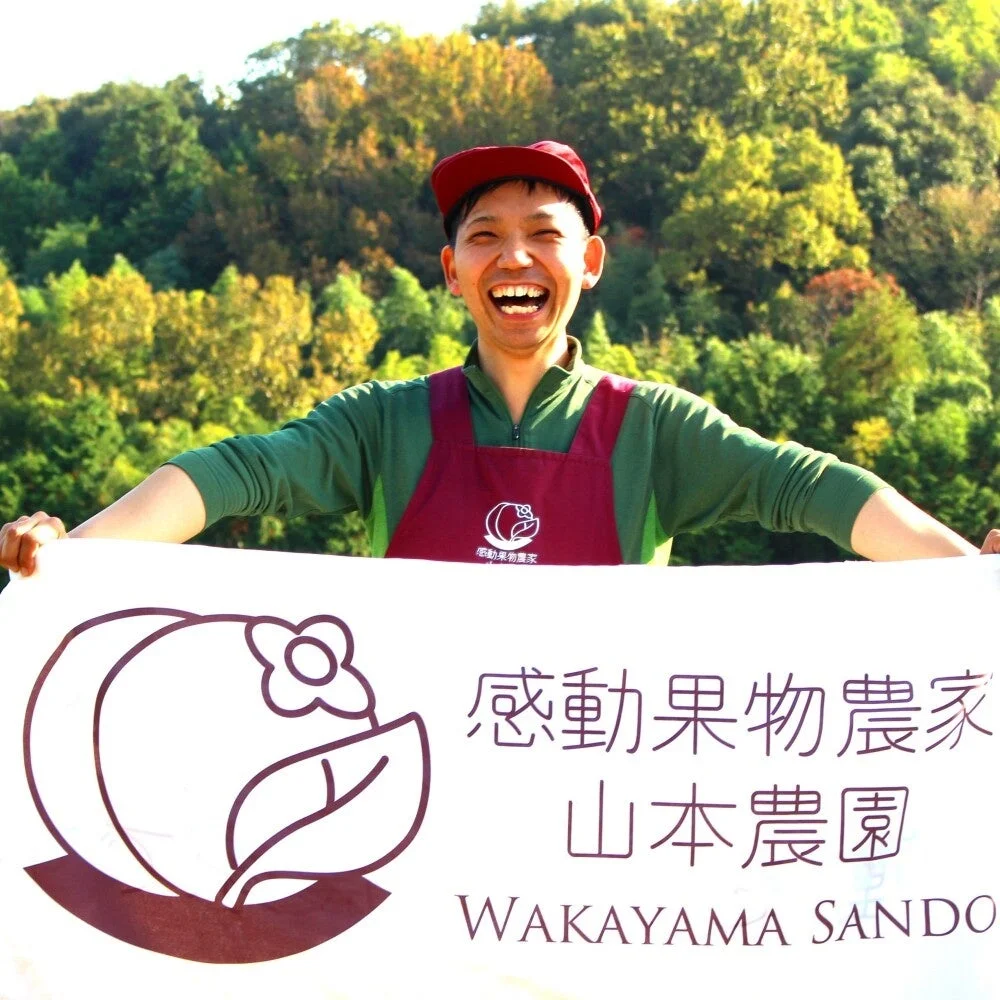 赤い帽子とエプロンを着用した男性が、緑豊かな背景の前で満面の笑みを浮かべています