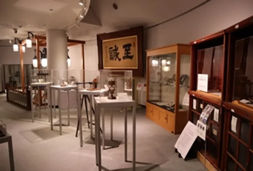 博物館の展示室で、ガラスケースや台座に様々な展示品が並べられている様子。壁には「誠至」と書かれた大きな額が飾られており、歴史や文化に関する展示であることがうかがえる。