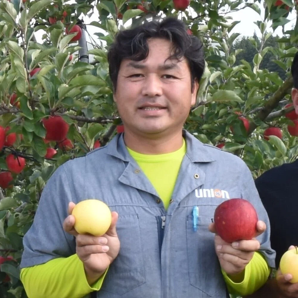 リンゴの木を背景に、笑顔の男性が赤いリンゴと黄色いリンゴをそれぞれ手に持っています