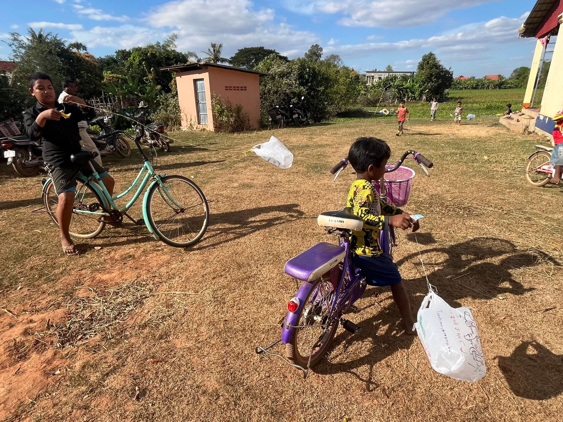 晴れた屋外で子供たちが遊ぶ様子。少年たちが自転車に乗り、プラスチック袋を凧のように飛ばして楽しんでいます。