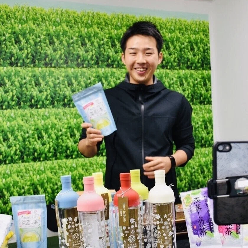 笑顔の男性が深蒸し茶のパッケージとカラフルな水出し茶ボトルを手に持ち、商品を紹介しています