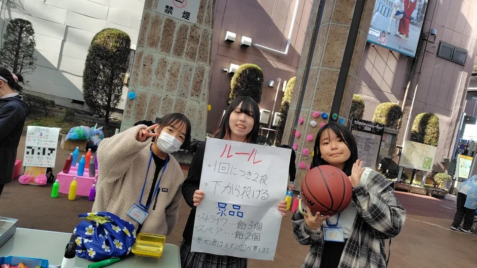 屋外のイベントで、3人の若い女性がゲームブースを運営しています。中央の女性はゲームのルールと景品が書かれたボードを持ち、右の女性はバスケットボールを抱え、左の女性はピースサインをしています。楽しげな雰囲気が伝わる一枚です。