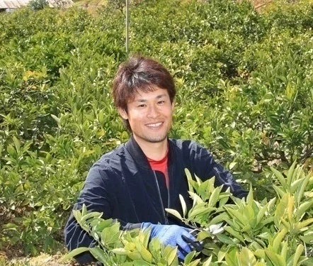 緑豊かな畑の中で、笑顔の男性が作業服と手袋を着用して植物の手入れをしている様子です