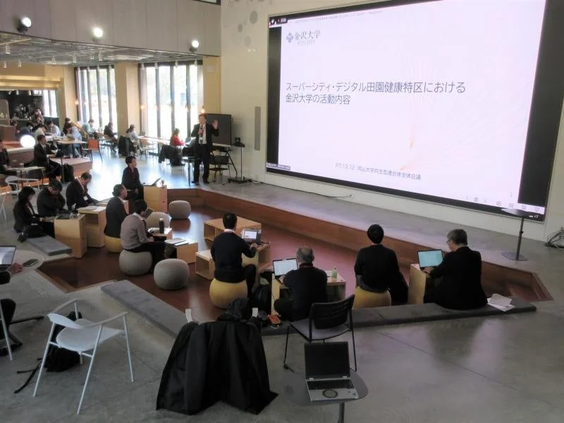 現代的な会場で、一人の男性が大型スクリーンに映し出された資料を使いプレゼンテーションを行っています。