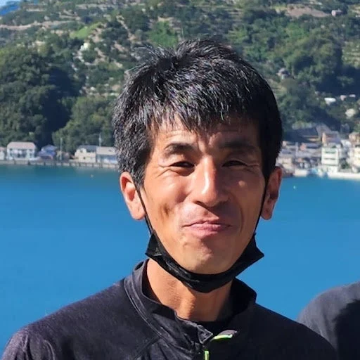 青い海と緑豊かな山々を背景に、マスクを顎に下げてにこやかに笑うアジア人男性のポートレート