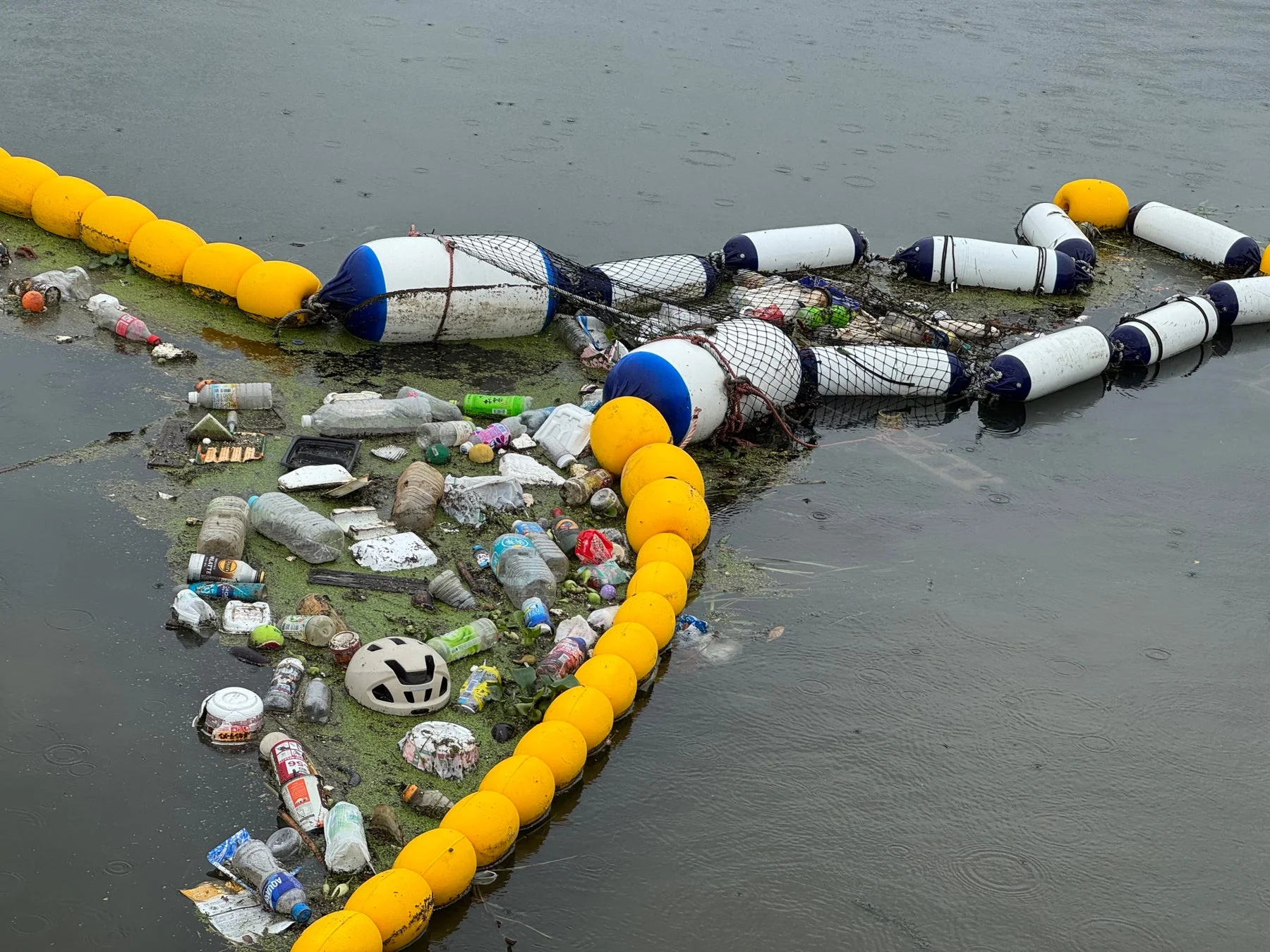 大量のプラスチックごみや様々な廃棄物が、黄色と白のブイによってせき止められ、水面に浮かんでいます