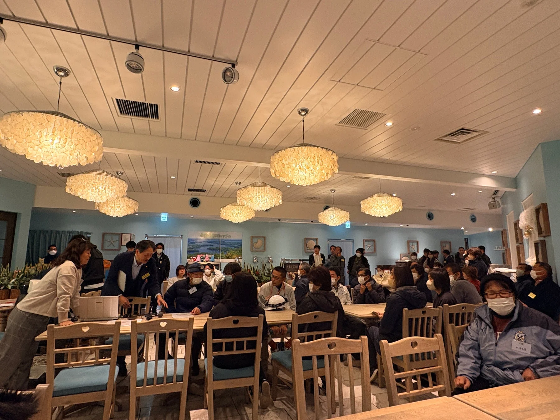 明るい屋内のカフェまたはレストランで、多くの人々がマスクを着用して集まっている様子