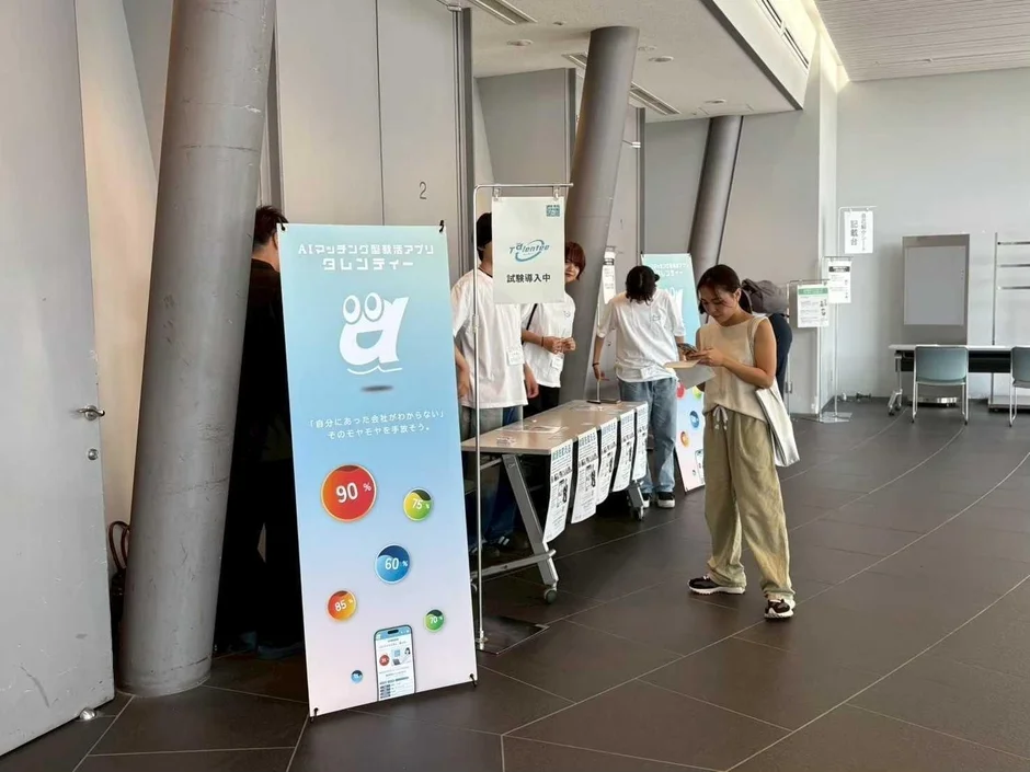 AIマッチング転職アプリ「タレンティー」のプロモーションブースが写っています。会場には数人が集まり、一人の女性がスマートフォンを操作しながらブースの案内を見ています。アプリの魅力が示された看板が目を引く、情報収集の場です。
