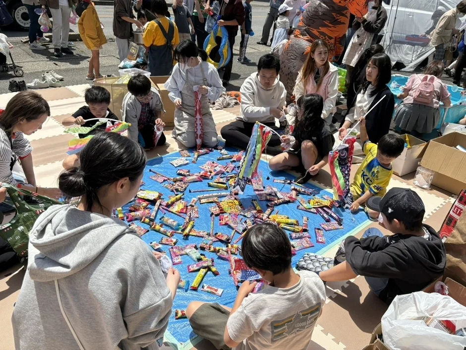 屋外のイベントで、多くの子供と大人が青いシートを囲んで座り、お菓子や鯉のぼり作りに興じている様子です。和やかな雰囲気が伝わってきます。